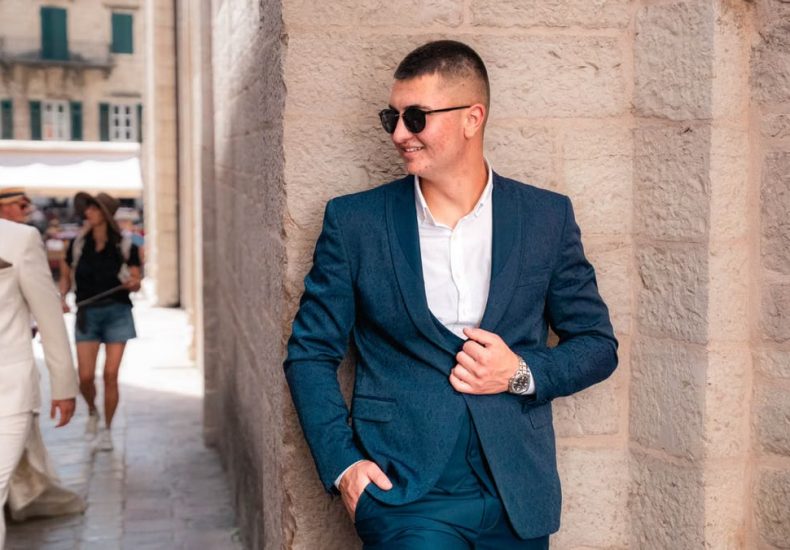 Confident man in a blue suit leans against a stone wall in charming Kotor, Montenegro.
